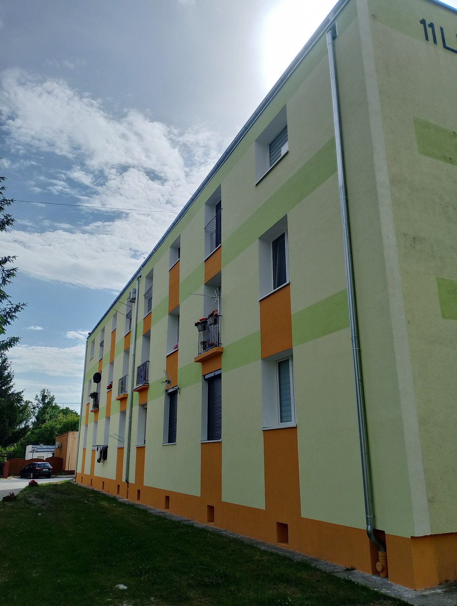 Odnowiona elewacja bloku z balkonami, pomalowana w poziome pasy: żółto-zielone i pomarańczowe. Widoczne okna, rynny i fragment trawnika. Słoneczny dzień.