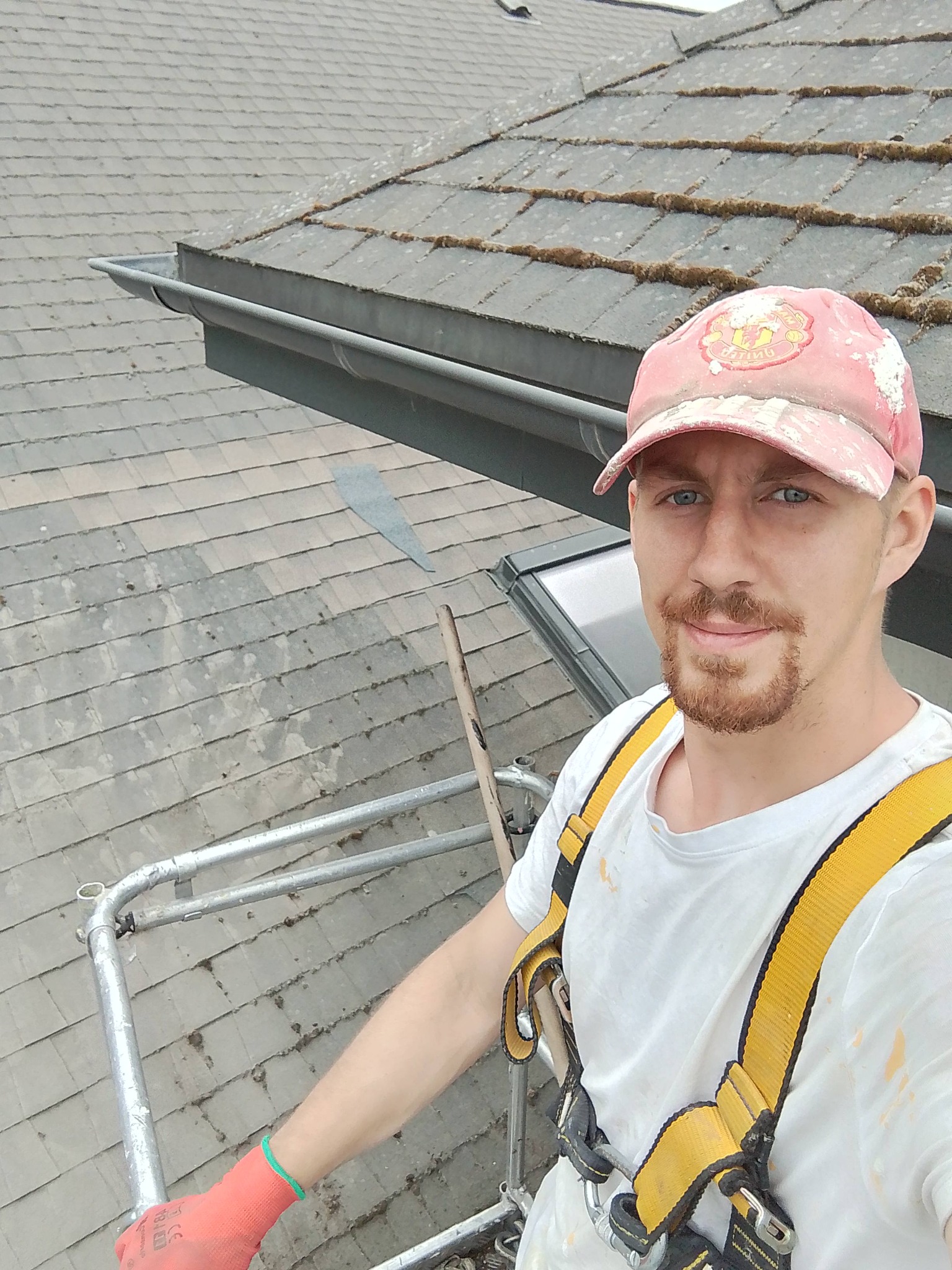 Pracownik na rusztowaniu, w uprzęży, na tle dachu pokrytego dachówką z mchem, w trakcie pracy. Widoczny fragment rynny i okno dachowe.