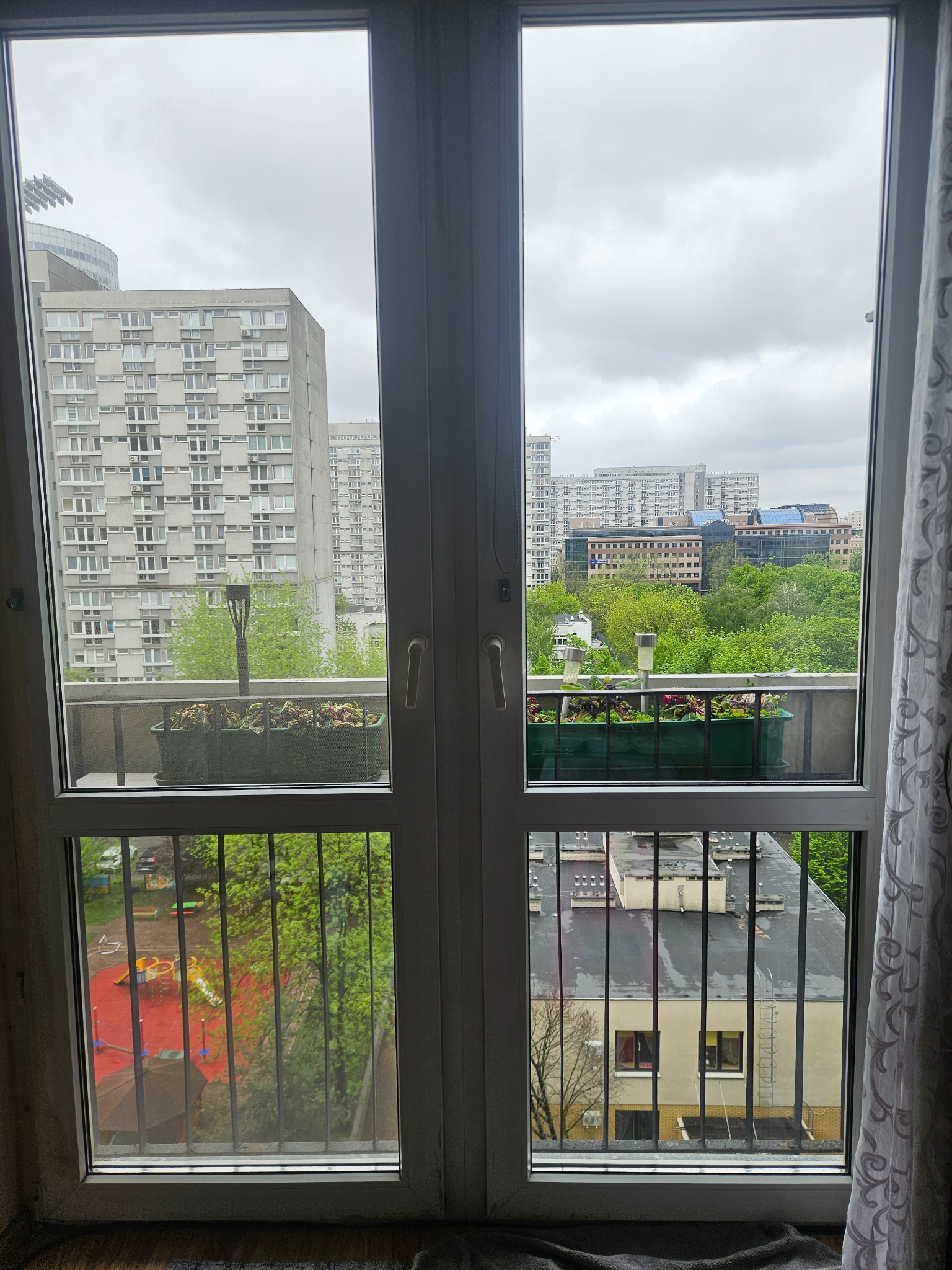 Widok z okna na blok, zieleń i plac zabaw. Na parapecie doniczki z kwiatami. Pochmurne niebo nad miastem.