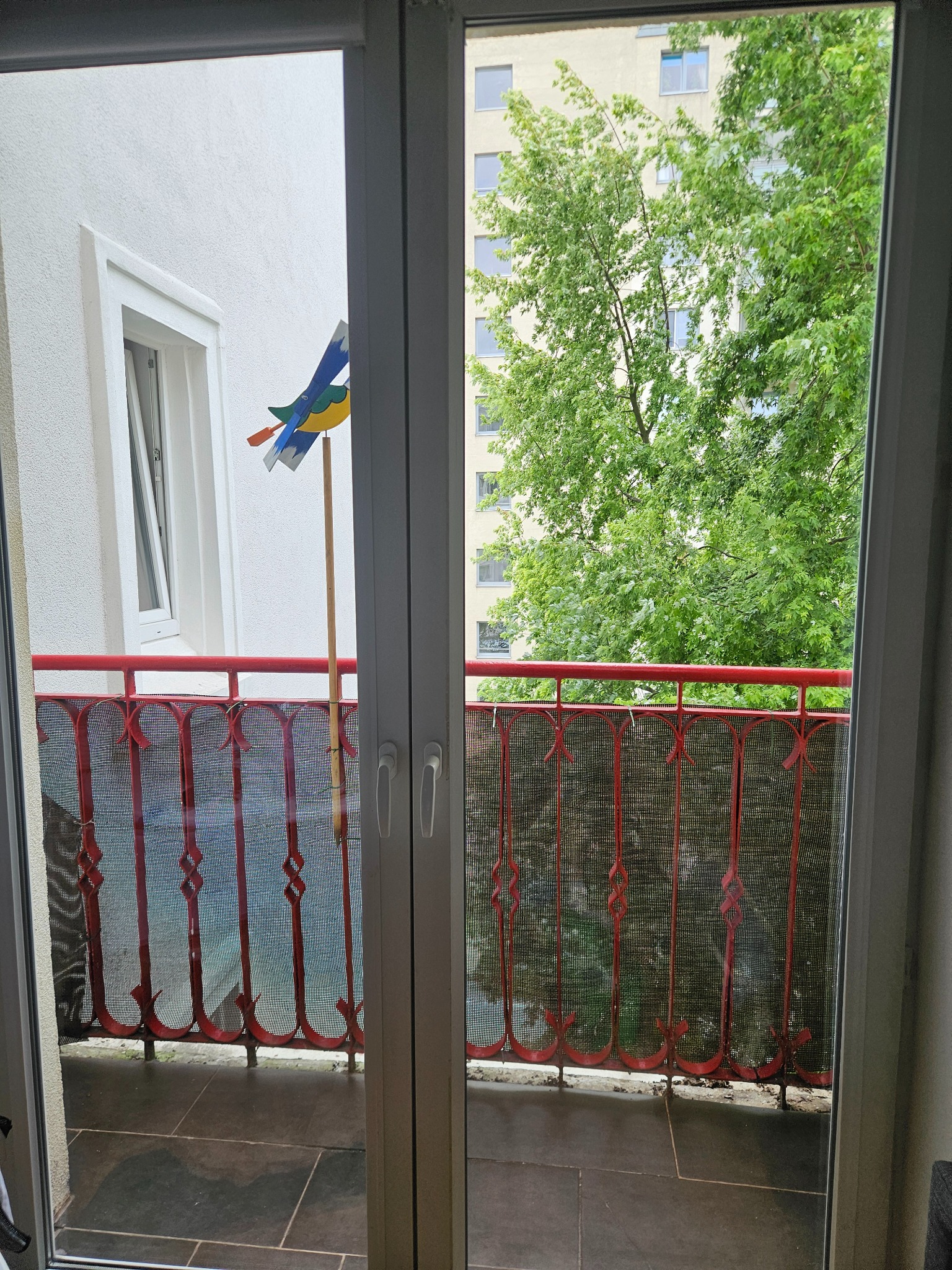 Widok z balkonu przez otwarte okno: czerwona balustrada, zabawka wiatrak, budynek i zieleń w tle. Ujęcie z wnętrza mieszkania.