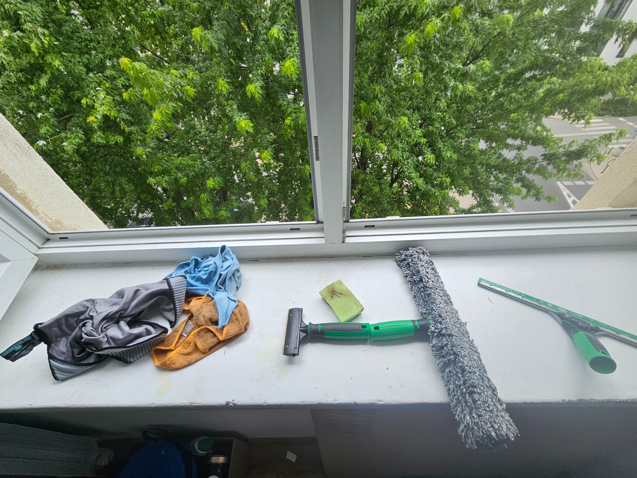 Narzędzia do mycia okien (ściągaczka, myjka, gąbka, szmatki) leżące na parapecie okna z widokiem na zielone drzewa. Sprzątanie po umyciu okien.