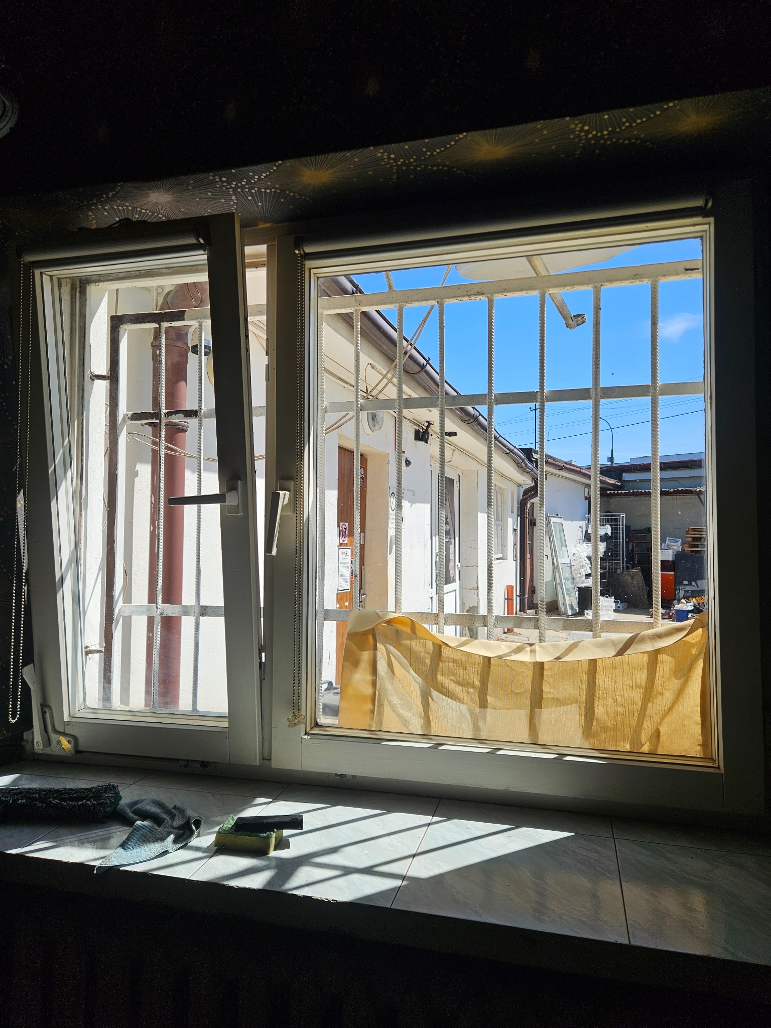 Otwarty, biały okno z kratami, przez które widać podwórko w słoneczny dzień. Na parapecie leżą narzędzia do czyszczenia szyb.