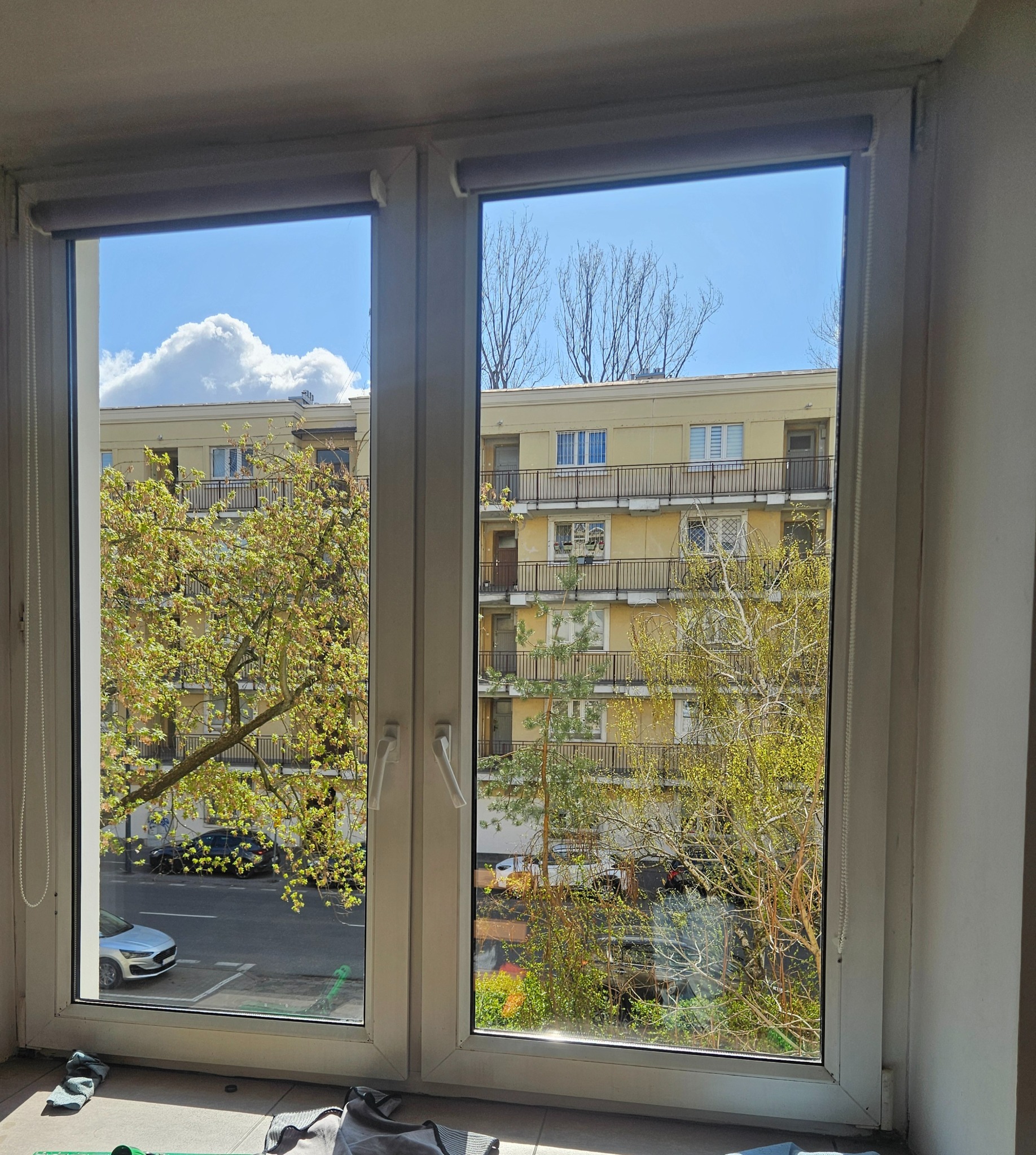 Dwuskrzydłowe, białe okno z widokiem na budynek i zieleń. Częściowo zasłonięte rolety. Na parapecie leżą ściereczki i zielony mop.