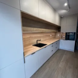 Nowoczesna kuchnia z zabudową w odcieniach szarości, drewnianym blatem i czarną armaturą, oświetlona punktowymi lampami sufitowymi.