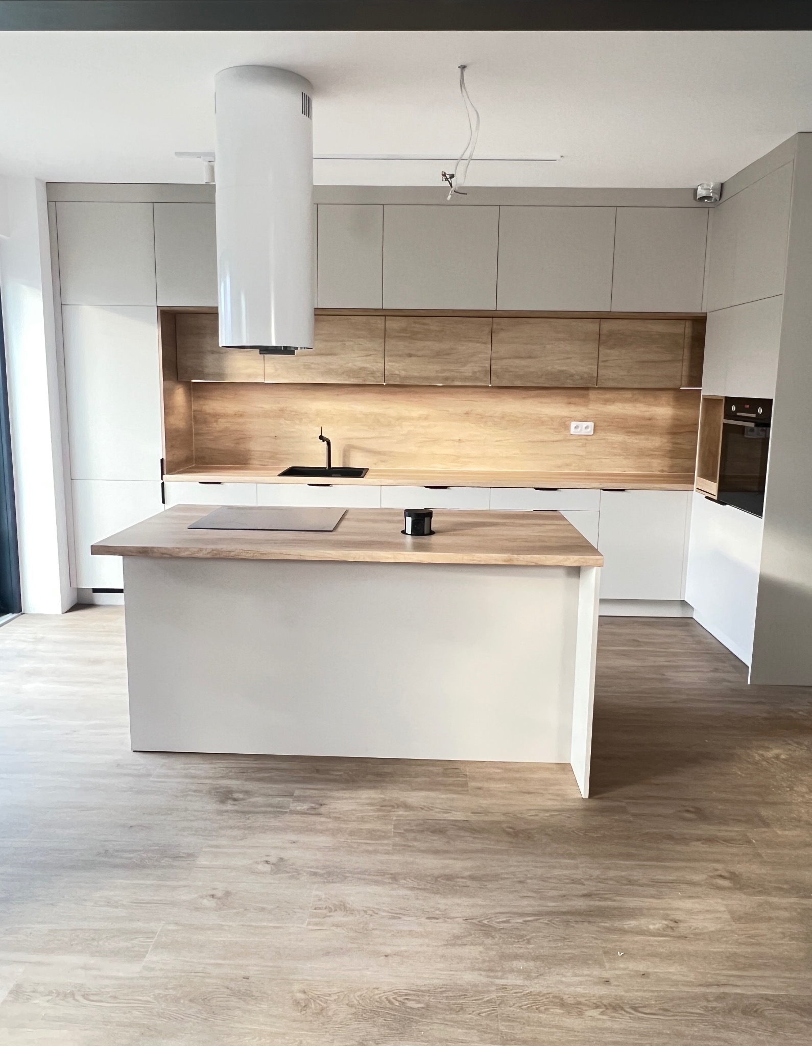 Nowoczesna kuchnia z wyspą, białe szafki i drewniane panele ścienne, cylindryczny okap, czarna armatura, płyta indukcyjna na wyspie.