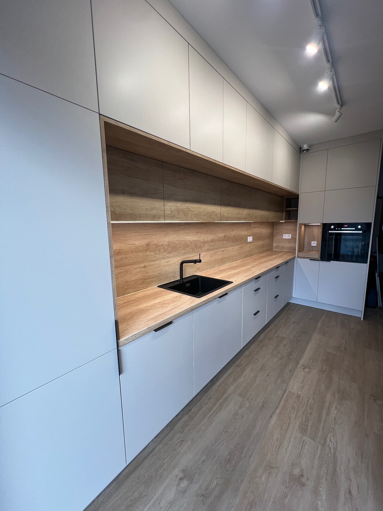 Nowoczesna kuchnia z zabudową w odcieniach szarości, drewnianym blatem i czarną armaturą, oświetlona punktowymi lampami sufitowymi.