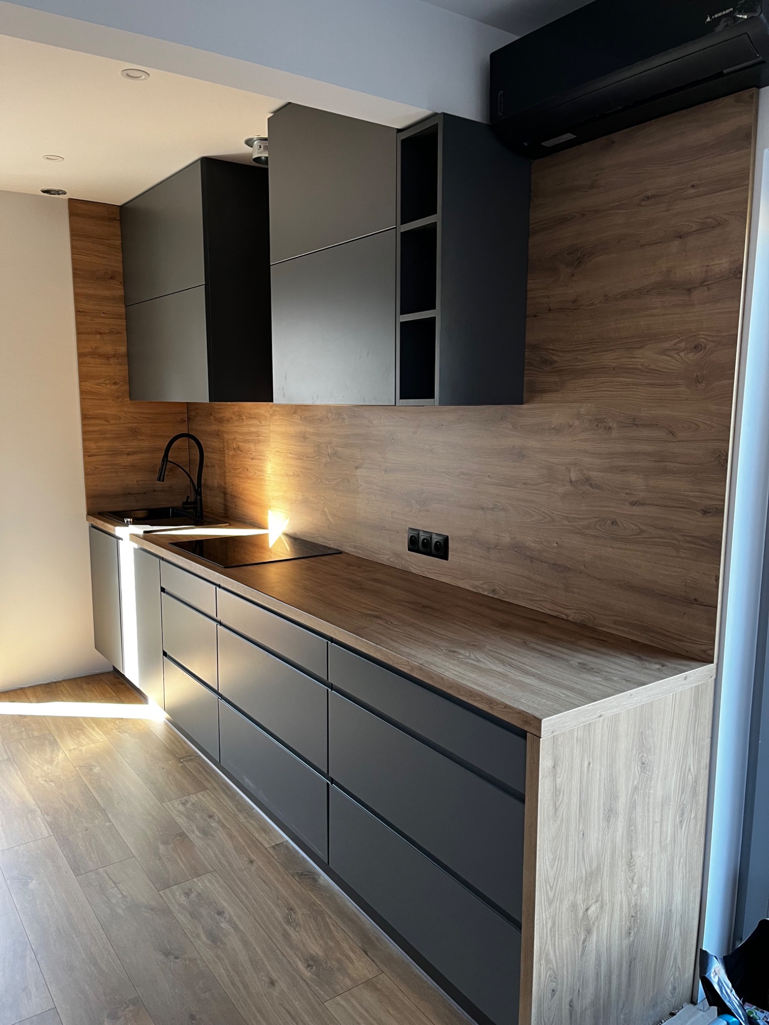 Nowoczesna, minimalistyczna kuchnia z szarymi frontami szafek i blatem imitującym drewno, czarna armatura i płyta indukcyjna, drewniana okładzina ścienna, oświetlenie punktowe i klimatyzator.