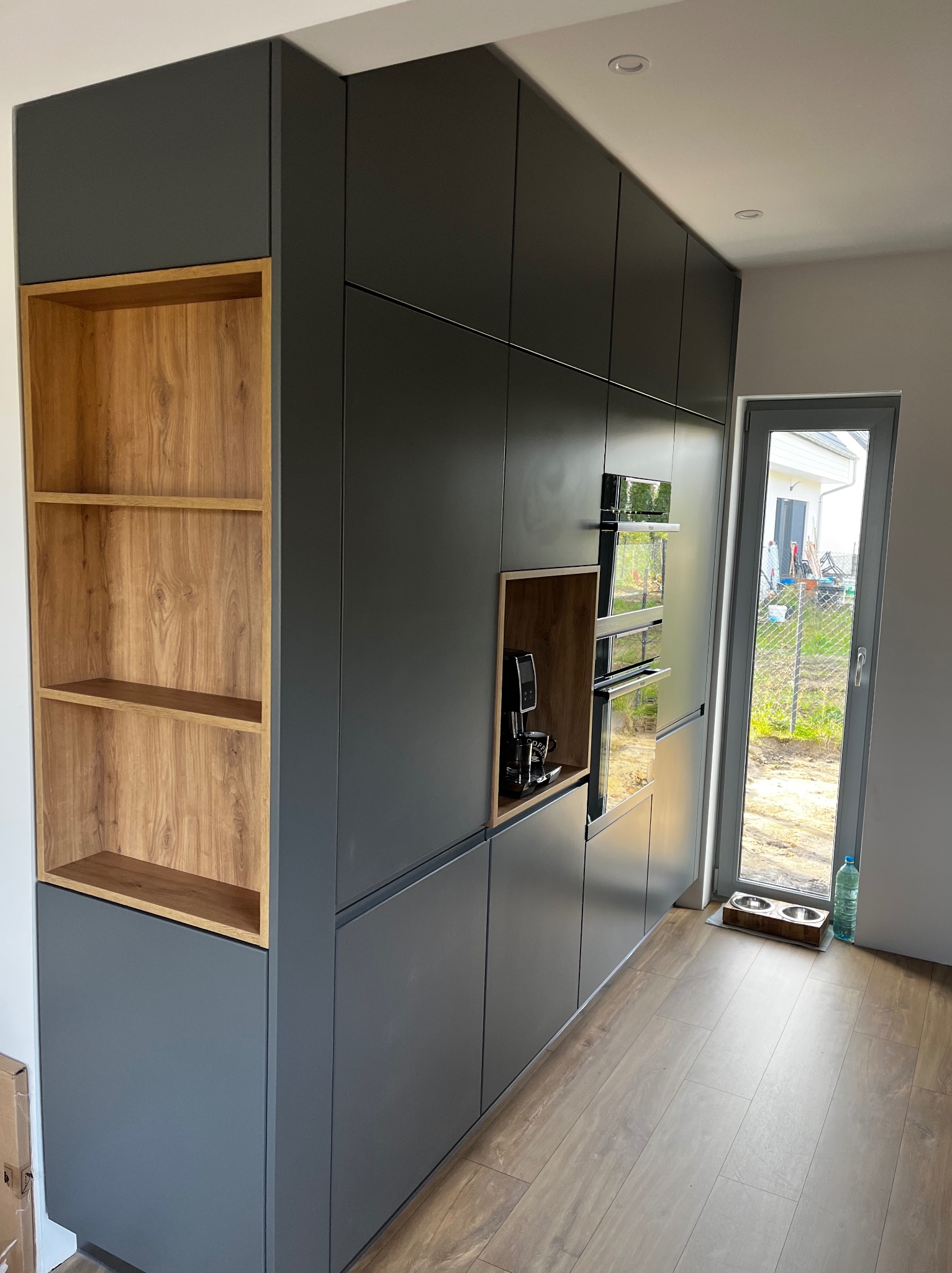 Nowoczesna zabudowa kuchenna w kolorze grafitowym z drewnianymi akcentami, wnęką z półkami i wbudowanymi piekarnikami, z widocznym ekspresem do kawy i podłogą imitującą drewno.