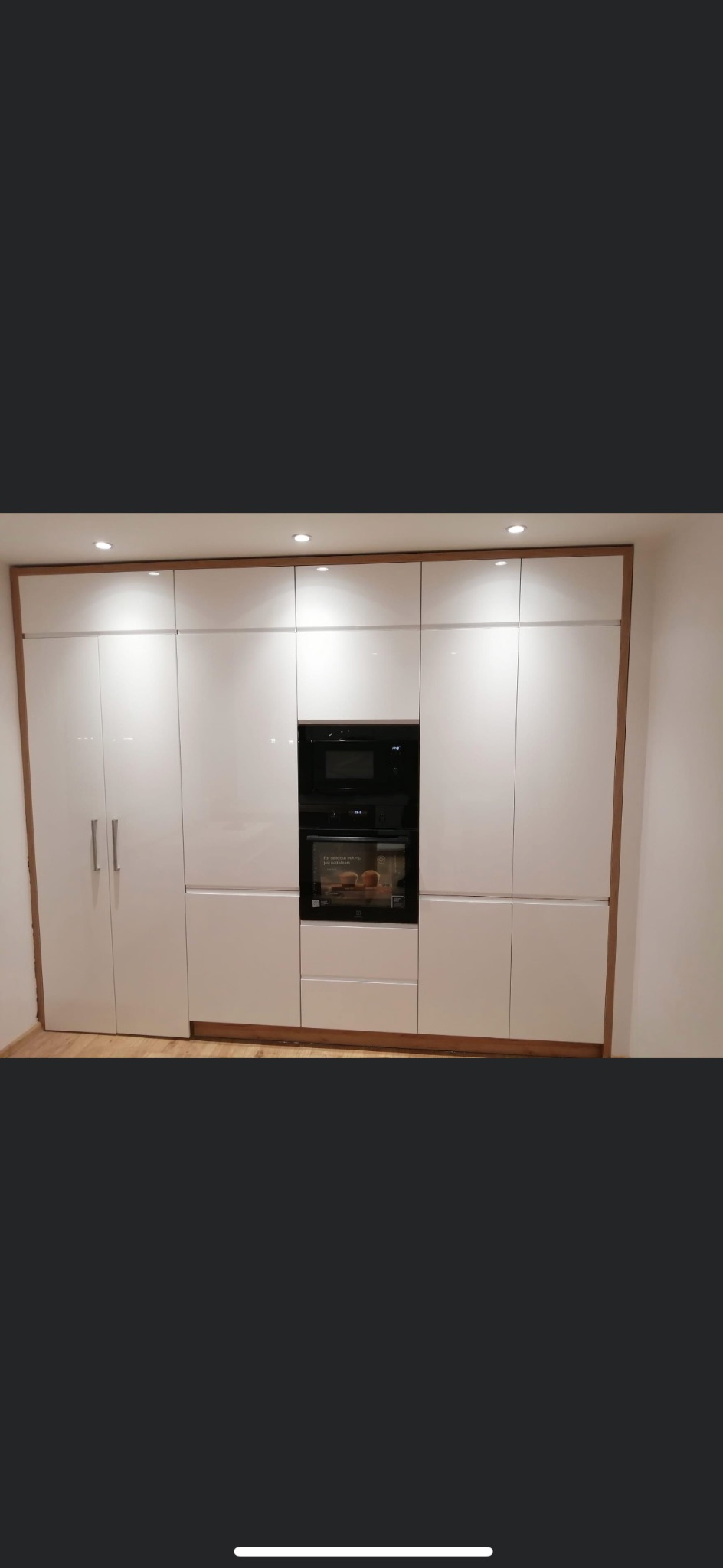 Białe, lakierowane szafki kuchenne z drewnianym obramowaniem i wbudowanym piekarnikiem i mikrofalówką.