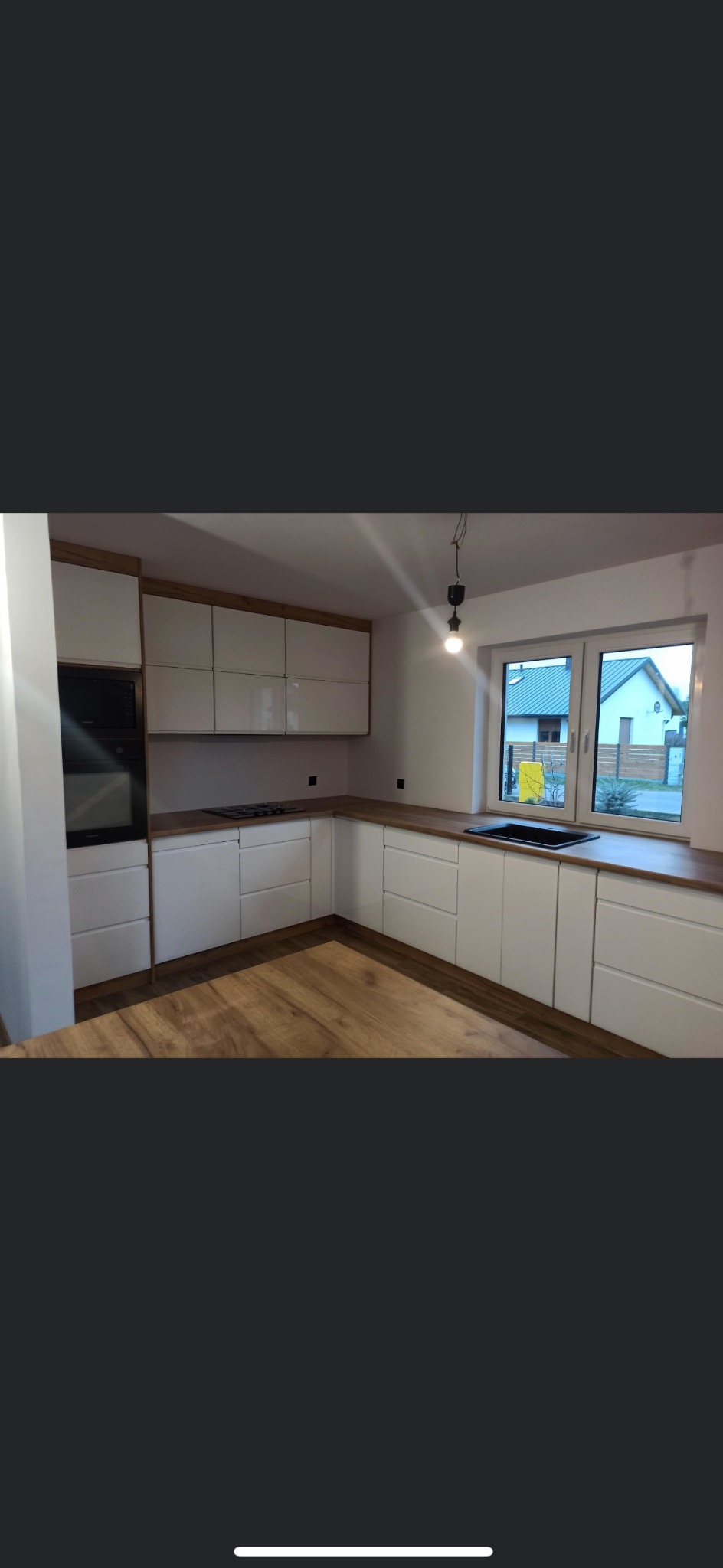 Nowoczesna, jasna kuchnia z białymi szafkami i drewnianymi blatami, oświetlona pojedynczą żarówką, widok na okno z domem w tle.