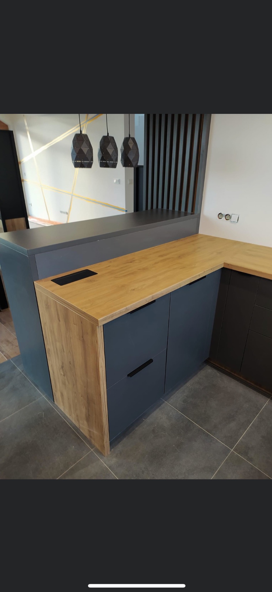 Nowoczesna kuchnia na wymiar z drewnianym blatem i szarymi frontami szafek, widoczne czarne uchwyty, trzy geometryczne lampy wiszące i dekoracyjne linie na ścianie w tle.