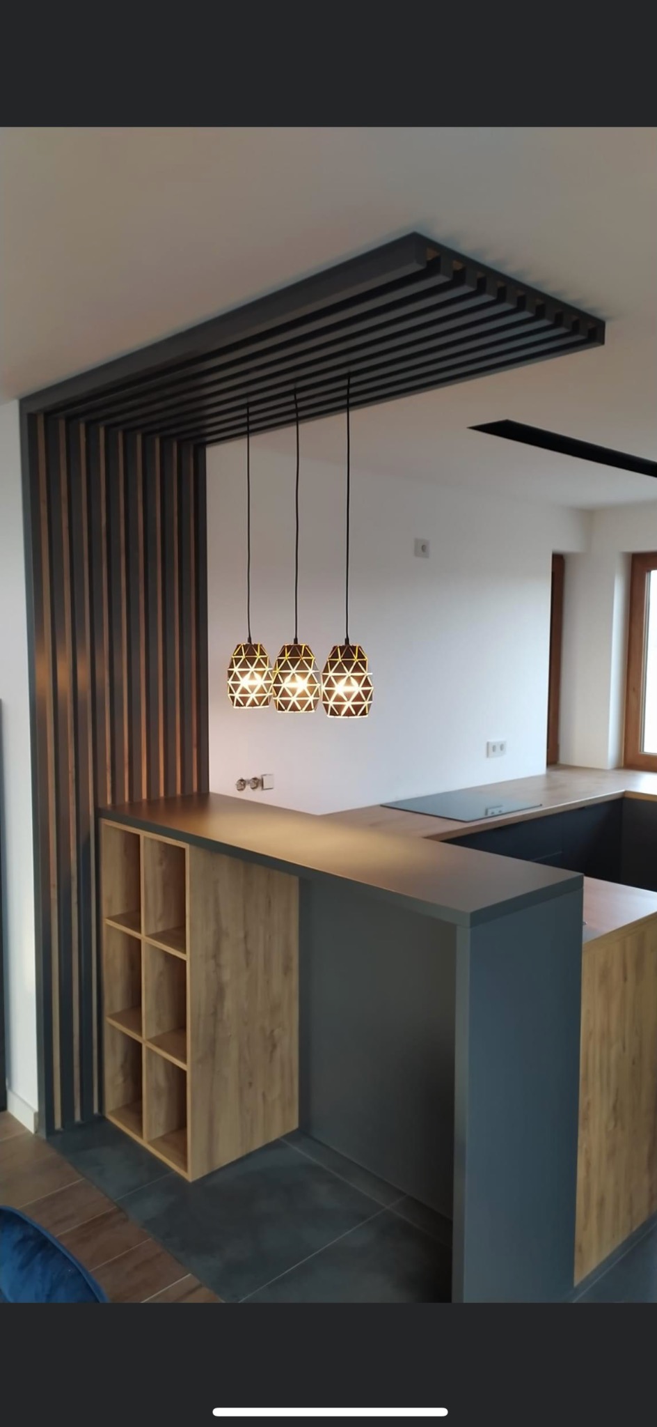Nowoczesna kuchnia z wyspą oddzieloną od salonu pionowymi lamelami i drewnianą zabudową z półkami, oświetlona trzema geometrycznymi lampami wiszącymi.