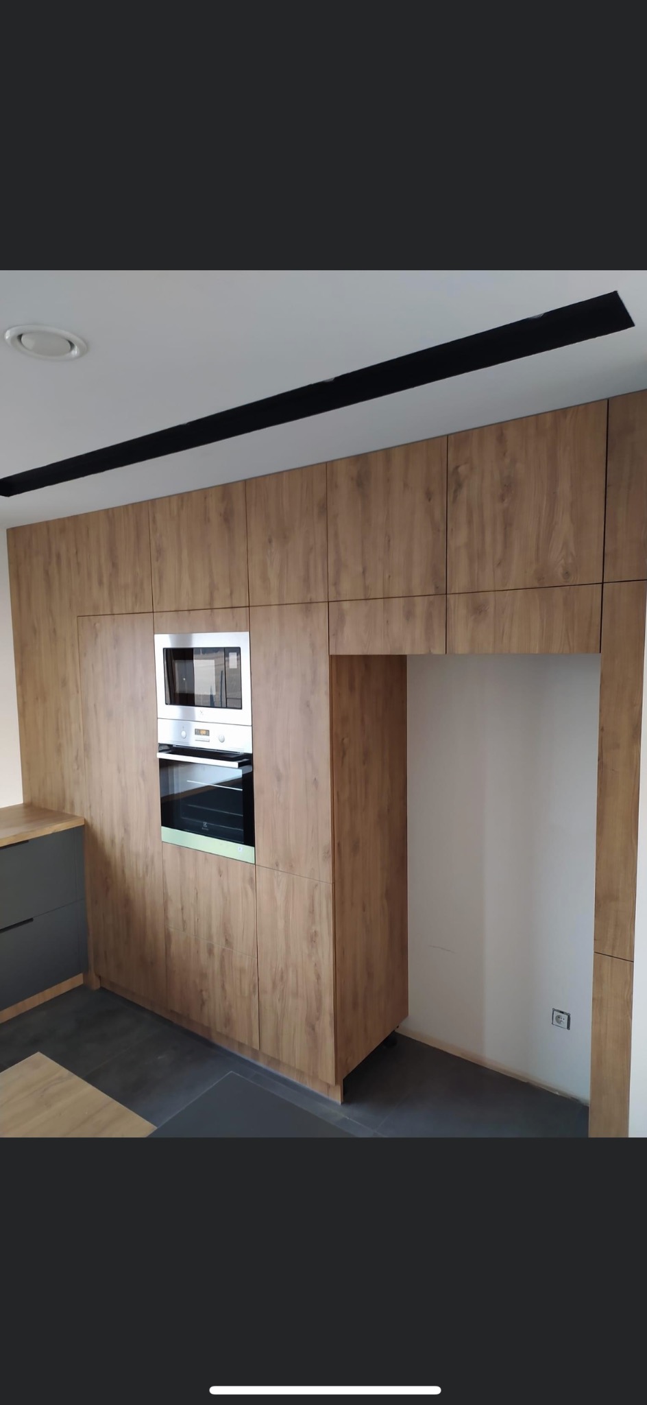 Drewnopodobne szafki kuchenne z wbudowanym piekarnikiem i mikrofalówką, czarny pas na suficie, szare blaty i ciemna podłoga.