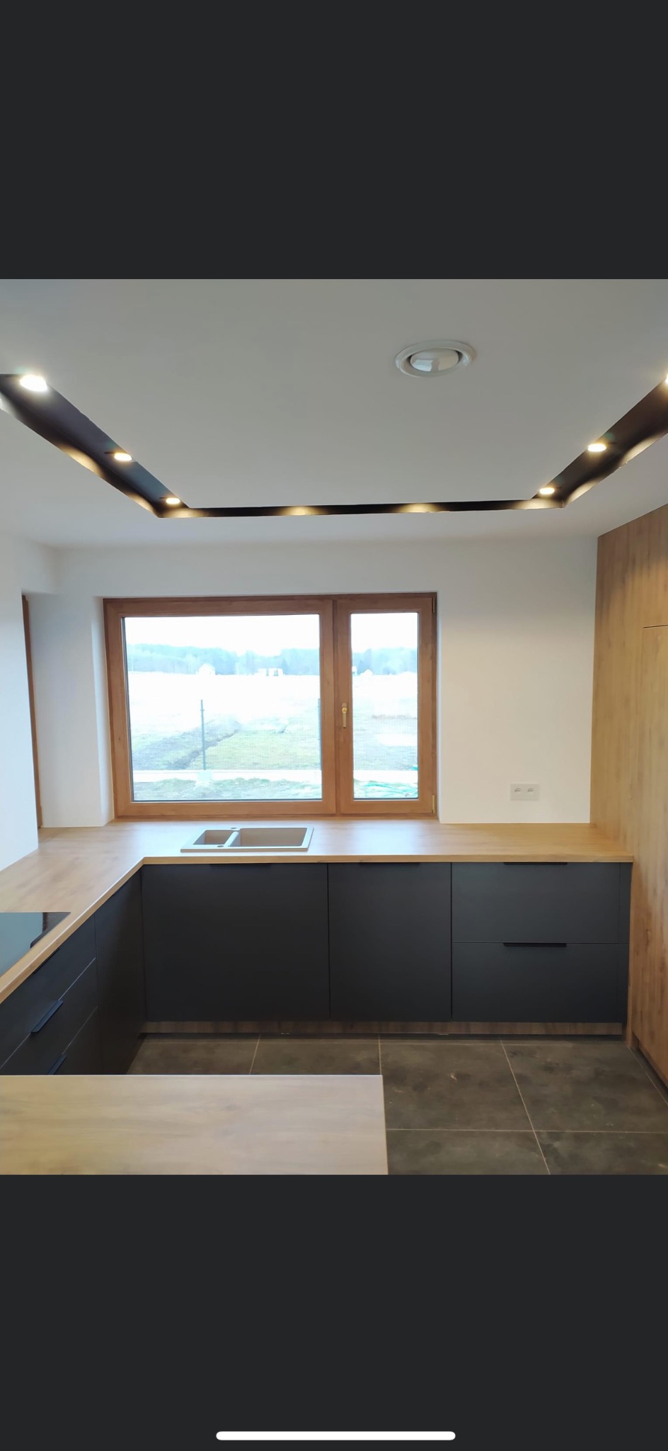 Nowoczesna kuchnia z czarnymi szafkami, drewnianym blatem i oknem z widokiem na zewnątrz, oświetlenie sufitowe LED