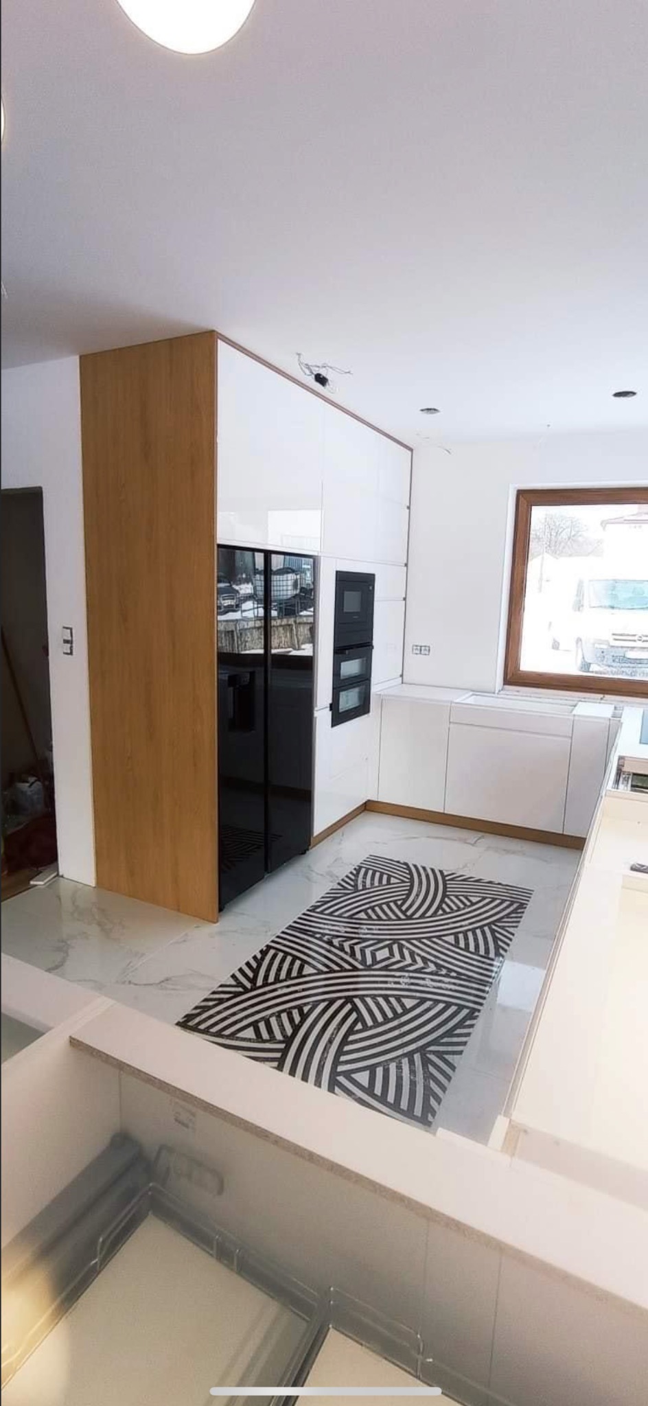 Nowoczesna kuchnia z białymi szafkami, drewnianą zabudową lodówki i czarnym dywanem w geometryczne wzory na marmurowej podłodze.