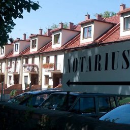 KANCELARIA NOTARIALNA RENATA MIKULEWICZ Notariusz - Szyld z napisem 'Notariusz' i strzałką, umieszczony przed szeregiem domów z czerwonymi dachami i balkonami z kwiatami, częściowo zasłonięty przez zaparkowane samochody.