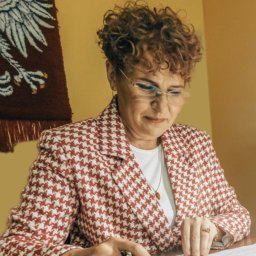 KANCELARIA NOTARIALNA RENATA MIKULEWICZ Notariusz - Kobieta w okularach i żakiecie w pepitkę, siedząca przy biurku i czytająca dokument, w tle godło Polski.