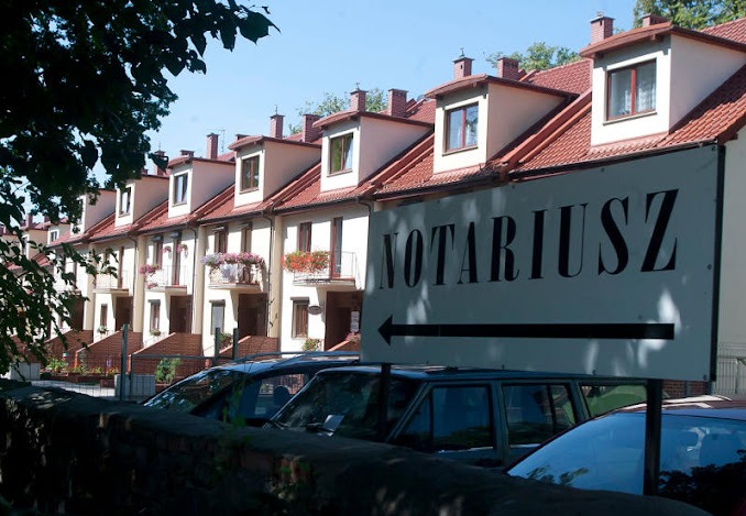 Szyld z napisem 'Notariusz' i strzałką, umieszczony przed szeregiem domów z czerwonymi dachami i balkonami z kwiatami, częściowo zasłonięty przez zaparkowane samochody.