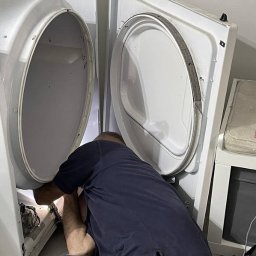 AGDKlinika - Mężczyzna w ciemnej koszulce naprawia pralkę automatyczną, zaglądając do wnętrza urządzenia. Widoczne otwarte drzwiczki i narzędzia.