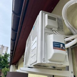 AGDKlinika - Biały klimatyzator na ścianie budynku z brązowym dachem, zamontowany na metalowym wsporniku. Widoczny budynek w tle i naklejka z adresem AGDKlinika.pl.