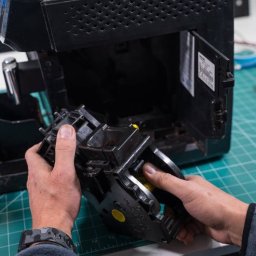 AGDKlinika - Demontaż modułu zaparzającego w ekspresie do kawy. Widoczne ręce serwisanta, wnętrze urządzenia i blat warsztatowy z podziałką.