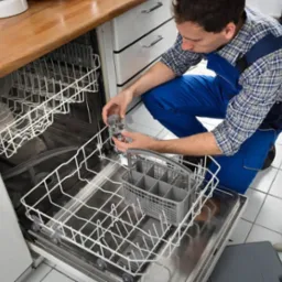 Mężczyzna w niebieskim kombinezonie naprawia zmywarkę, trzymając filtr. Na podłodze leżą rękawice i klucze.