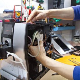 AGDKlinika - Naprawa ekspresu do kawy: ręce serwisanta przy rozłożonym urządzeniu, widoczne elementy wewnętrzne, wężyki i narzędzia. W tle warsztat z miernikiem.