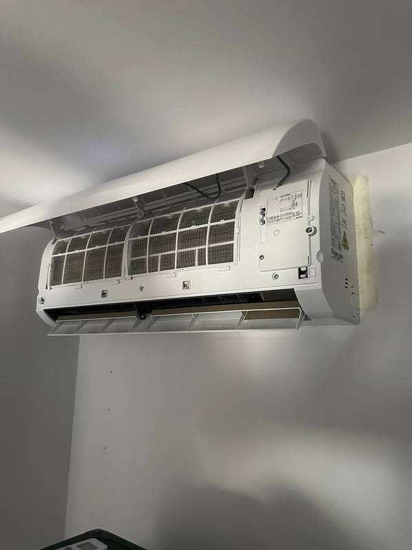 Biały klimatyzator ścienny z otwartą obudową, ukazujący filtry powietrza. Widoczne elementy montażowe i oznaczenia producenta. Ściana i sufit w tle.