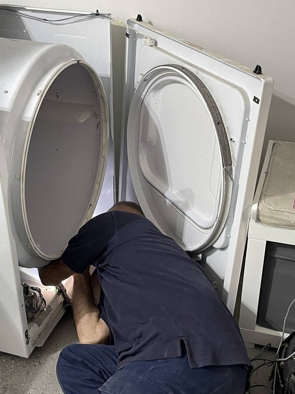 Mężczyzna w ciemnej koszulce naprawia pralkę automatyczną, zaglądając do wnętrza urządzenia. Widoczne otwarte drzwiczki i narzędzia.