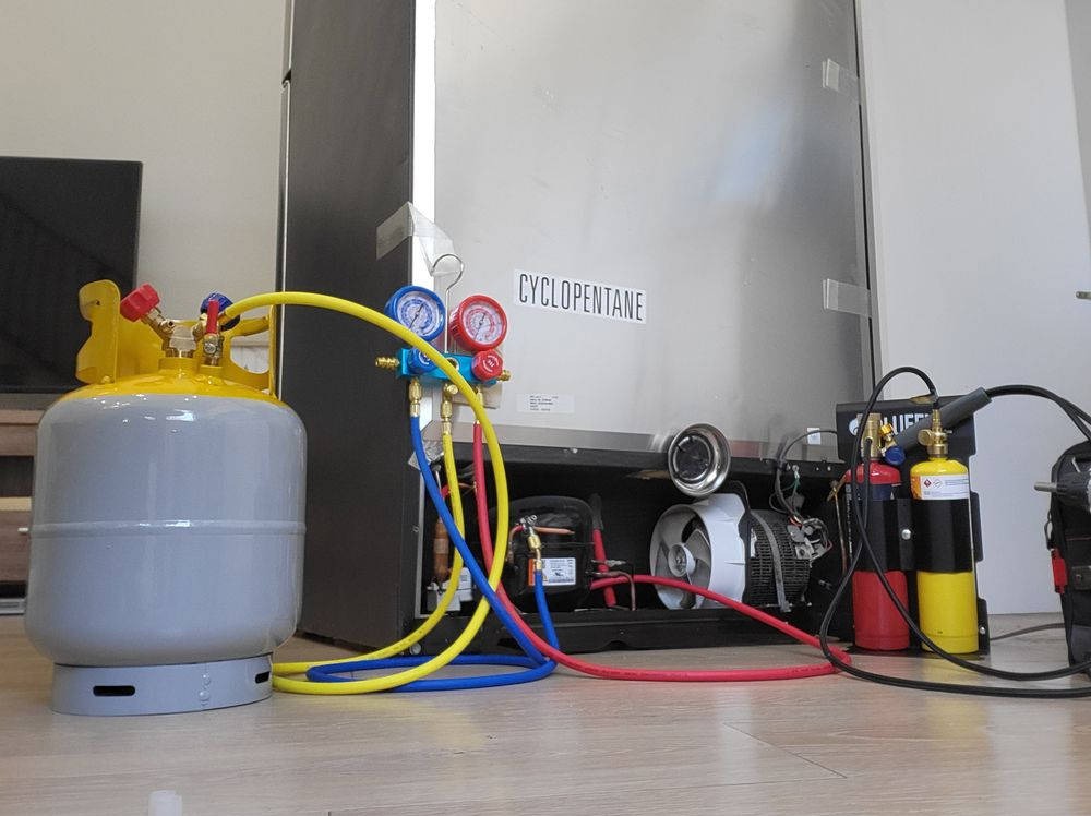 Butla z manometrami podłączona do lodówki podczas serwisu. Widoczne przewody i narzędzia serwisowe. Etykieta z napisem 'CYCLOPENTANE' na obudowie lodówki.