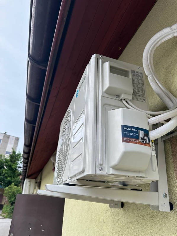 Biały klimatyzator na ścianie budynku z brązowym dachem, zamontowany na metalowym wsporniku. Widoczny budynek w tle i naklejka z adresem AGDKlinika.pl.