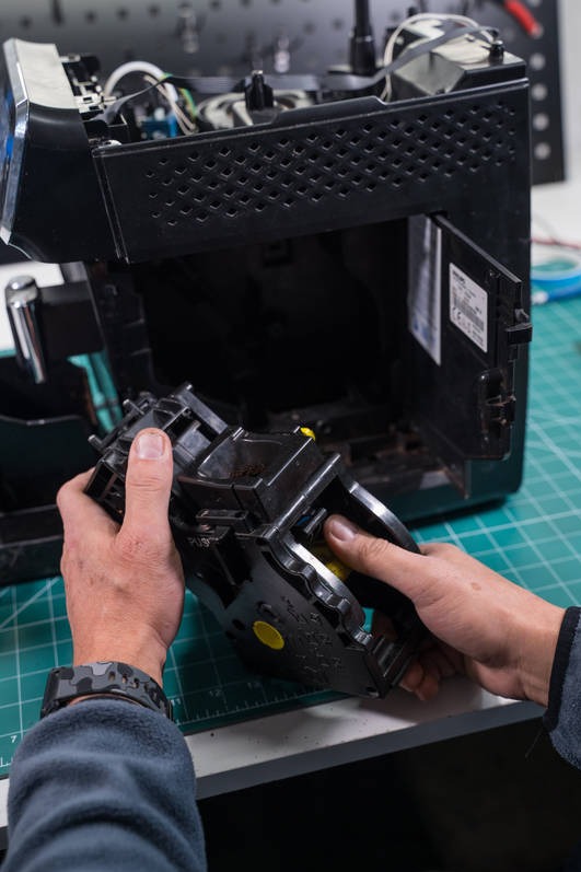 Demontaż modułu zaparzającego w ekspresie do kawy. Widoczne ręce serwisanta, wnętrze urządzenia i blat warsztatowy z podziałką.