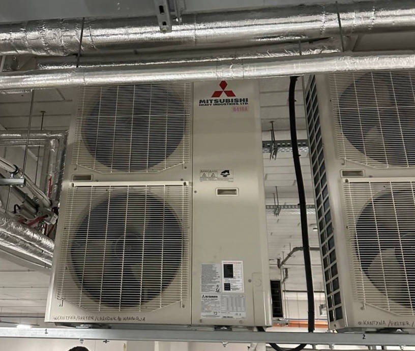 Białe, przemysłowe klimatyzatory Mitsubishi Heavy Industries LTD zamontowane pod sufitem w pomieszczeniu. Widoczne rury wentylacyjne.