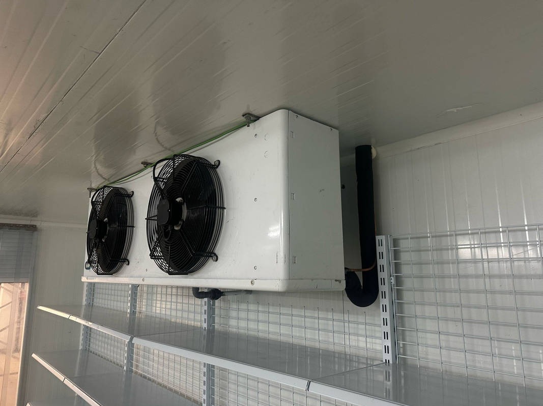 Biała przemysłowa chłodnica sufitowa z dwoma wentylatorami, rurami i metalowymi półkami w chłodni z panelami ściennymi.