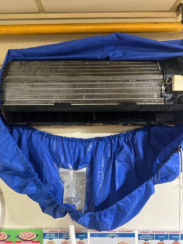 Klimatyzator zabezpieczony niebieską osłoną podczas czyszczenia. Widoczne żebra chłodnicy i biały wąż odprowadzający wodę. Tło stanowią reklamy pizzy.