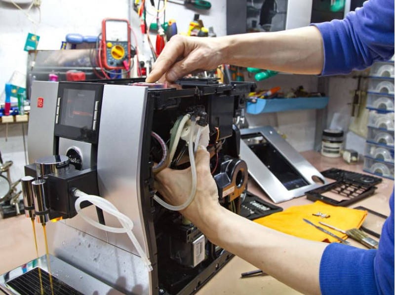 Naprawa ekspresu do kawy: ręce serwisanta przy rozłożonym urządzeniu, widoczne elementy wewnętrzne, wężyki i narzędzia. W tle warsztat z miernikiem.