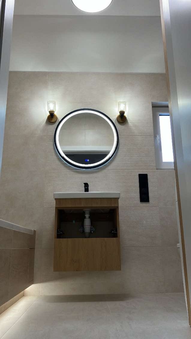 Nowoczesna łazienka z podświetlanym okrągłym lustrem, umywalką na szafce z drewnopodobnym frontem, i dwoma kinkietami ze szklanymi kloszami, oświetlona lampą sufitową