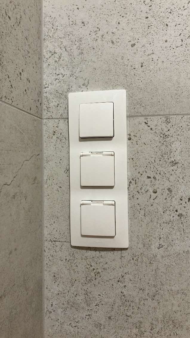 Potrójny, biały włącznik światła z klapkami, zamontowany na szarej, nakrapianej ścianie w łazience.