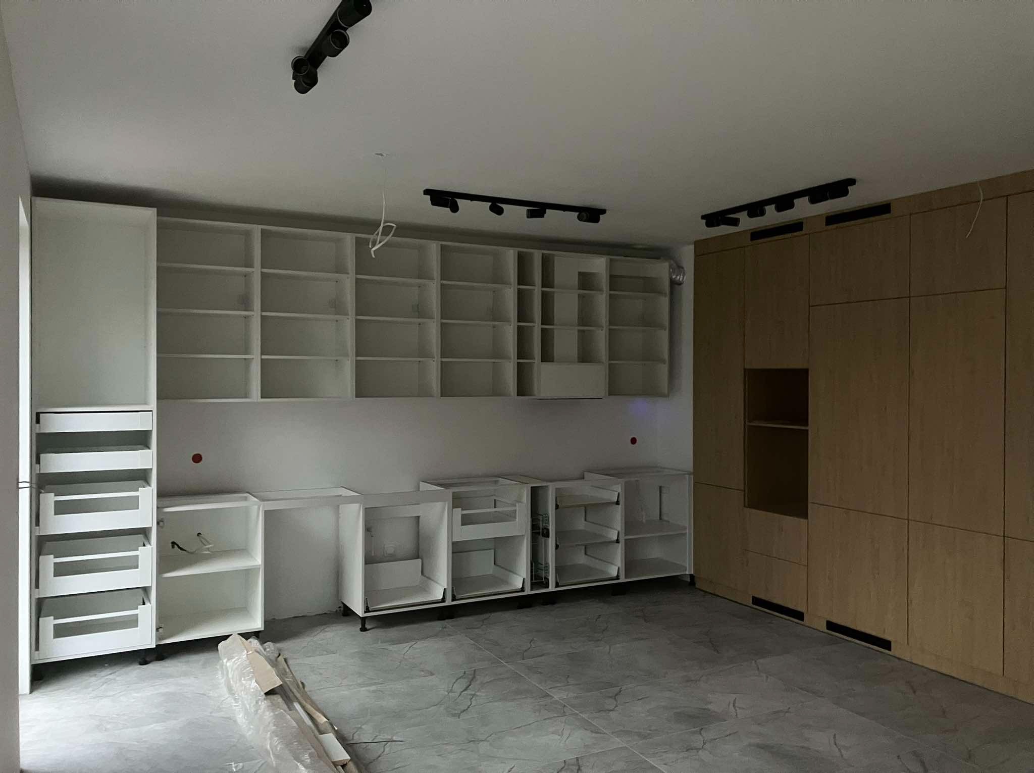 Wnętrze w trakcie montażu zabudowy kuchennej: widoczne białe i drewniane szafki bez frontów, szyny oświetleniowe na suficie, szare płytki podłogowe, elementy instalacji elektrycznej wystające ze...