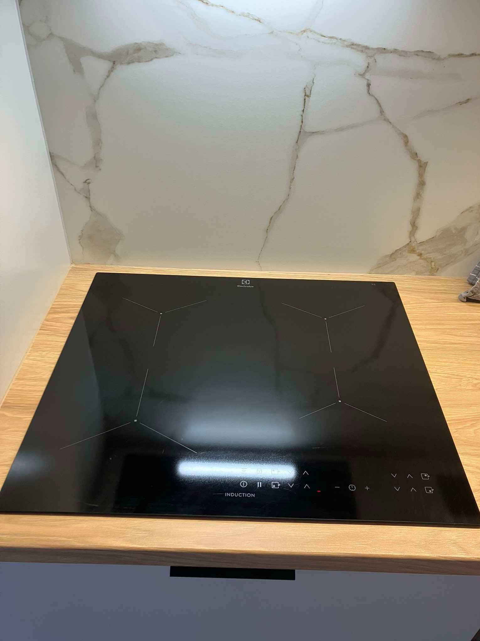 Czarna płyta indukcyjna Electrolux zamontowana w blacie z jasnego drewna, z panelem dotykowym i wzorem imitującym marmur na ścianie w tle.