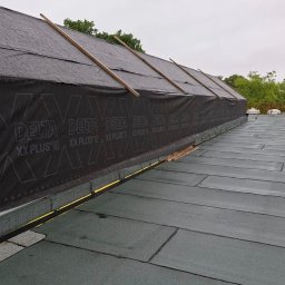 Dachy Płaskie Dawid Karpiak - Dach pokryty papą z widoczną membraną Delta XX Plus D, zabezpieczoną drewnianymi listwami. Widok z góry, fragment zieleni w tle.