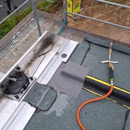 Dachy Płaskie Dawid Karpiak - Naprawa dachu z użyciem palnika dekarskiego i łaty bitumicznej. Widoczny fragment rynny. Prace dekarskie na płaskim dachu z papy.