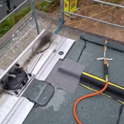 Naprawa dachu z użyciem palnika dekarskiego i łaty bitumicznej. Widoczny fragment rynny. Prace dekarskie na płaskim dachu z papy.