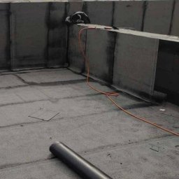 Dachy Płaskie Dawid Karpiak - Dekarz pracuje na płaskim dachu, widoczna rolka papy, zgrzewarka i pomarańczowy kabel. W tle budynki i pochmurne niebo.