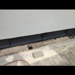 Dachy Płaskie Dawid Karpiak - Detal dachu przy ścianie budynku po uszczelnieniu. Widoczne elementy papy, cegła i kostka brukowa. Praca w trakcie realizacji, z bliska.