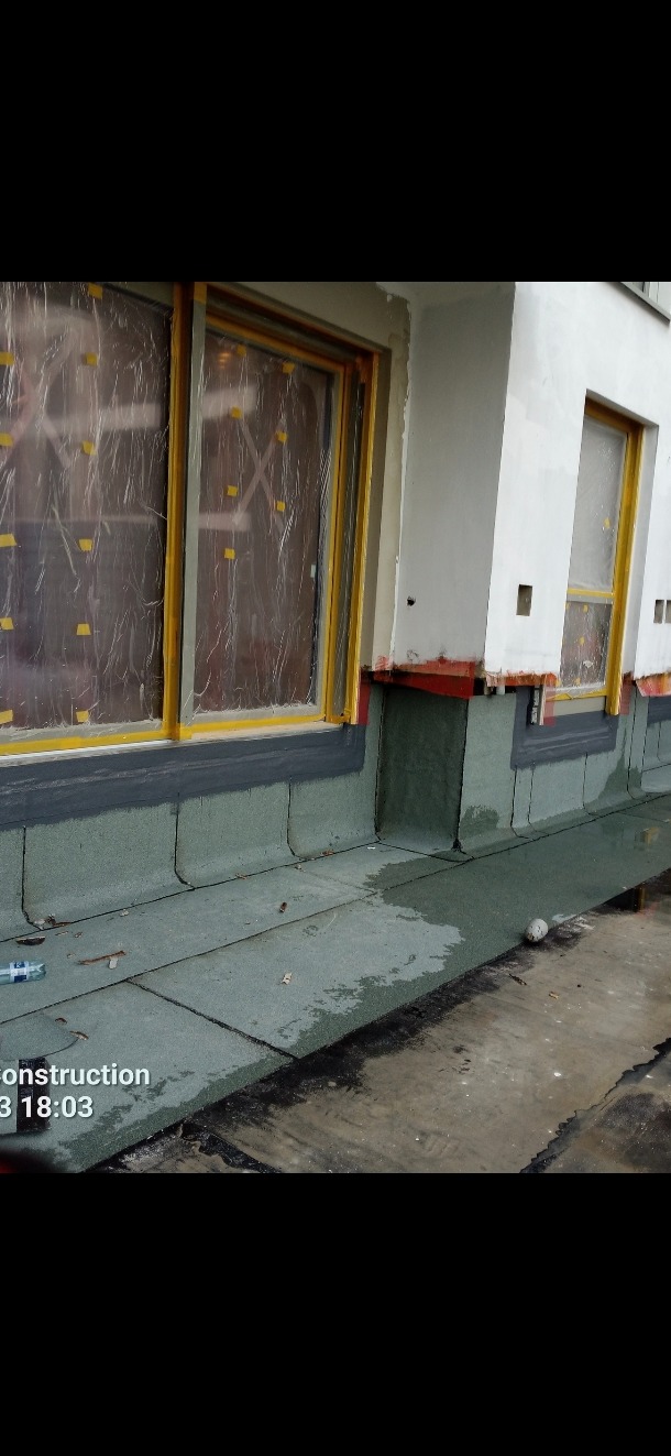 Prace dekarskie: zabezpieczone okna folią i żółtą taśmą, nowy zielony dach z papy bitumicznej przy ścianie budynku. Widoczny fragment orynnowania.