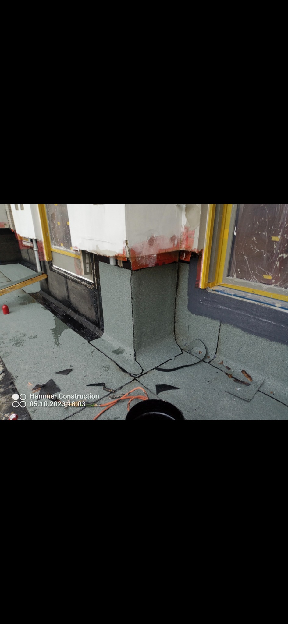 Detale krycia dachu papą termozgrzewalną wokół okien i komina. Widoczne narzędzia i odpływ. Szary kolor papy kontrastuje z czarnym wykończeniem.