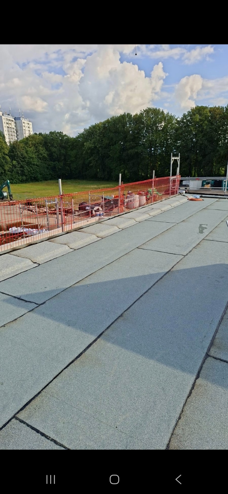 Wykończony dach z papy termozgrzewalnej, zabezpieczony barierkami. W tle budynek mieszkalny i zieleń. Widok z góry na płaski dach po remoncie.