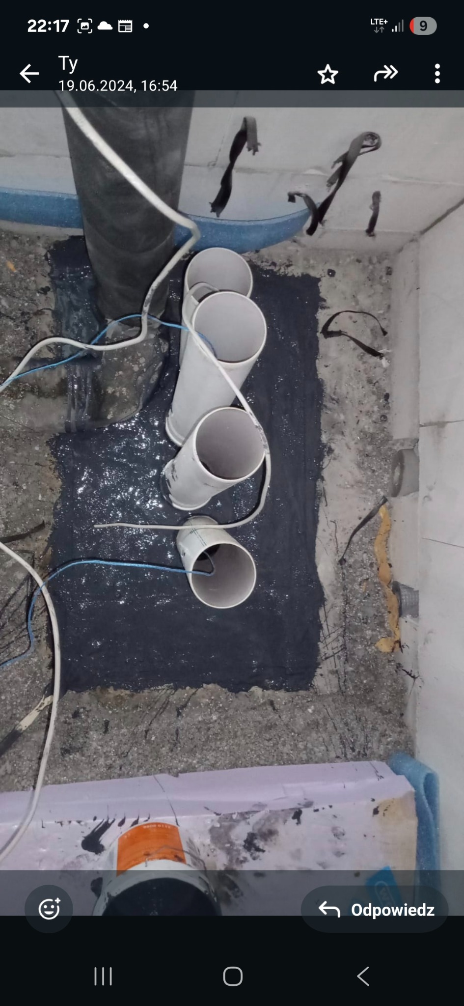 Hydroizolacja dachu płaskiego: cztery pionowe rury kanalizacyjne wystają z czarnej, gumowej powłoki hydroizolacyjnej na betonowej powierzchni dachu.