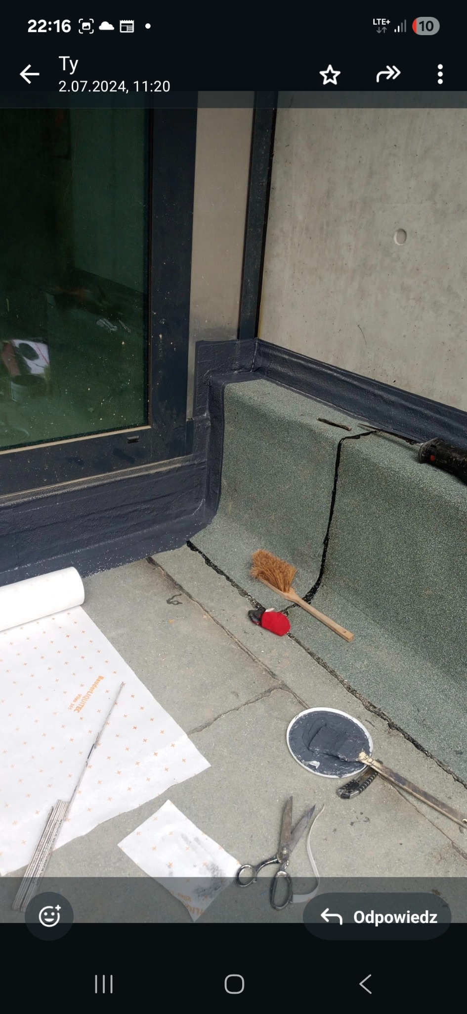 Szczegółowe ujęcie dachu płaskiego z papy termozgrzewalnej, uszczelnionego przy oknie. Widoczne narzędzia: szczotka, nożyczki, pędzel i zwój papy. Etap prac dekarskich.